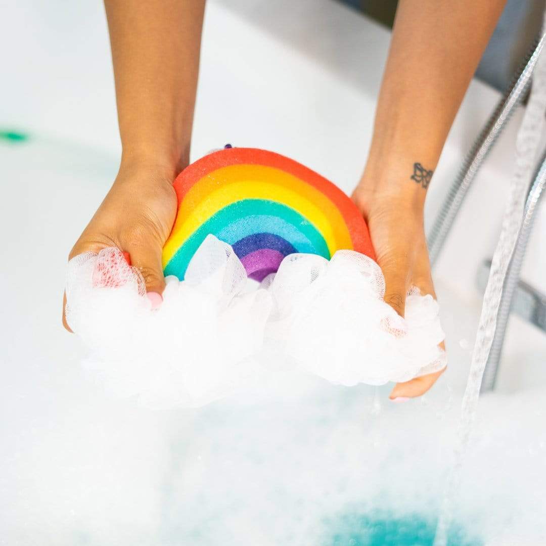 Hands holding Nectar Life Large Rainbow Loofah Bath Sponge over a sudsy bathtub, showing the colorful rainbow sponge lathering with white foam around the mesh loofah clouds
