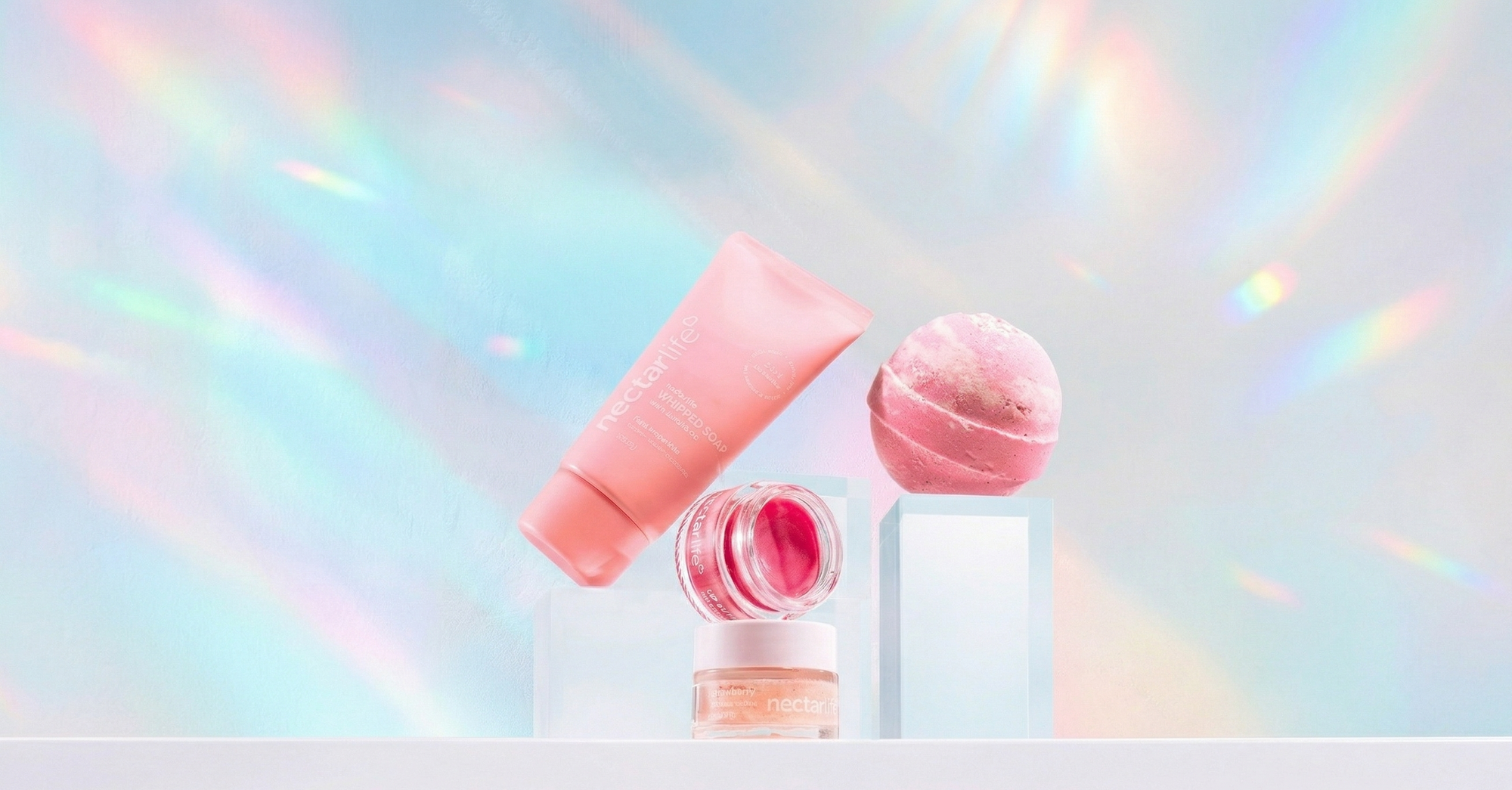 Three Nectar Life products styled on clear acrylic risers against a holographic rainbow background: a pink Whipped Soap tube, an open body butter jar, and a pink bath bomb.