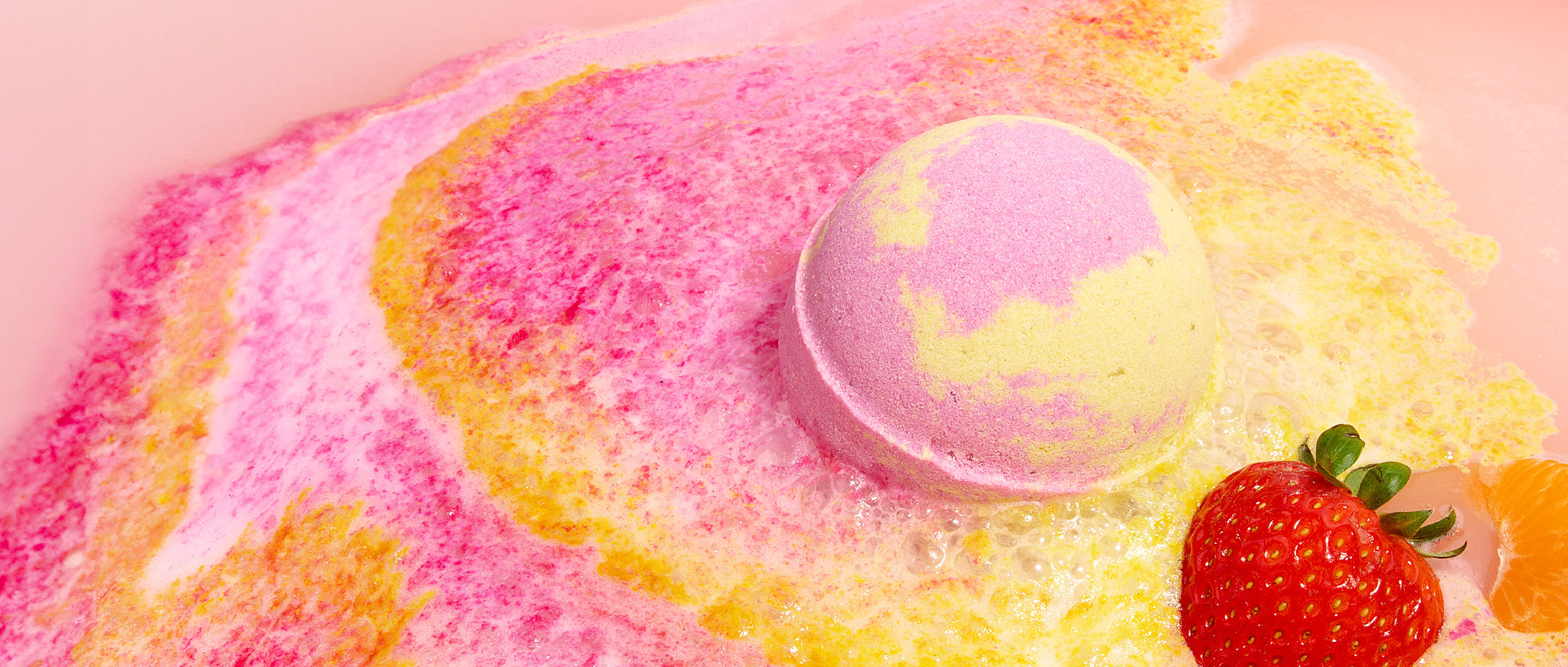 Fruit Smoothie Round Bath Bomb by Nectar Life fizzing in warm bath water, releasing swirls of pink, yellow, and orange foam with a fresh strawberry and tangerine segment floating alongside