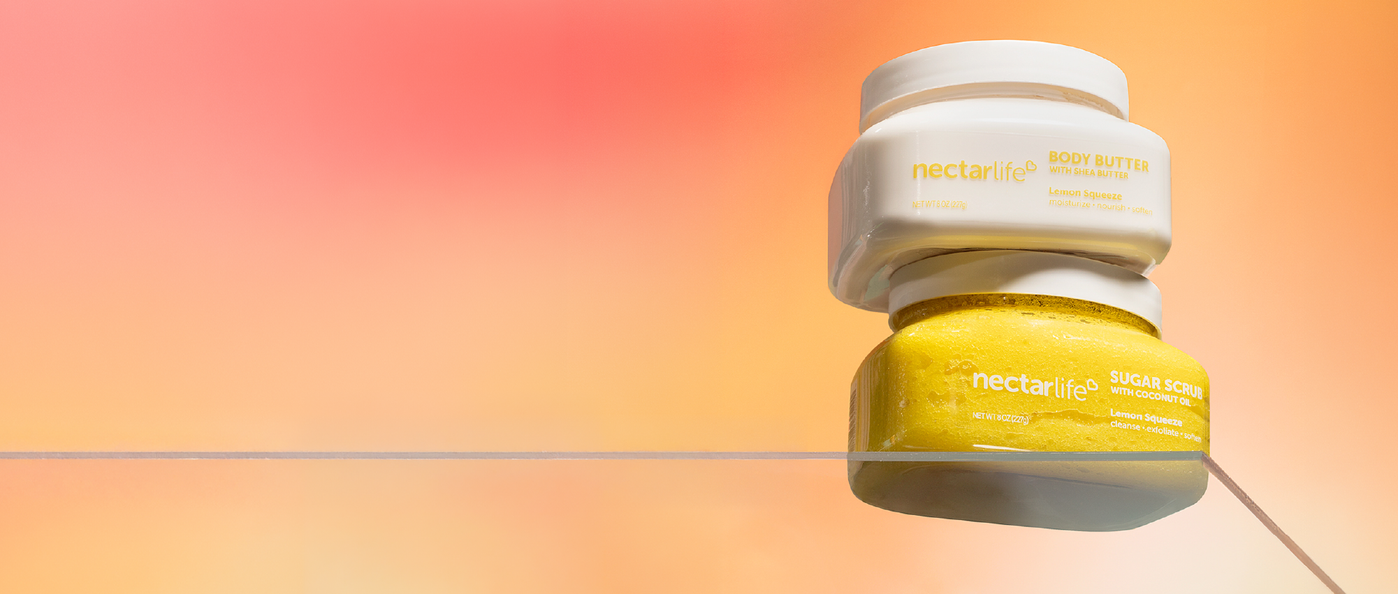 Two jars of Nectar Life Sugar Body Scrub and Body Butter with coconut oil in Lemon Squeeze Yellow stacked on a clear pedestal against a orange and coral background