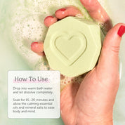 Hand holding a Nectar Life Essential Oil Benefit Bomb in warm fizzing water, clarifying soak with lavender eucalyptus and tea tree essential oils