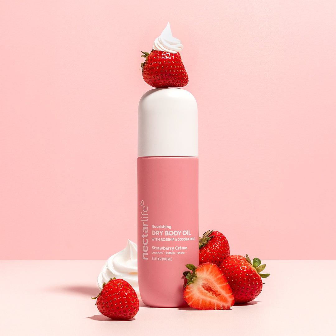 Strawberry Crème Nourishing Dry Body Oil by Nectar Life, pink spray bottle balanced on a fresh strawberry with whipped cream, styled with strawberries and cream on a pink background, 3.4 fl oz