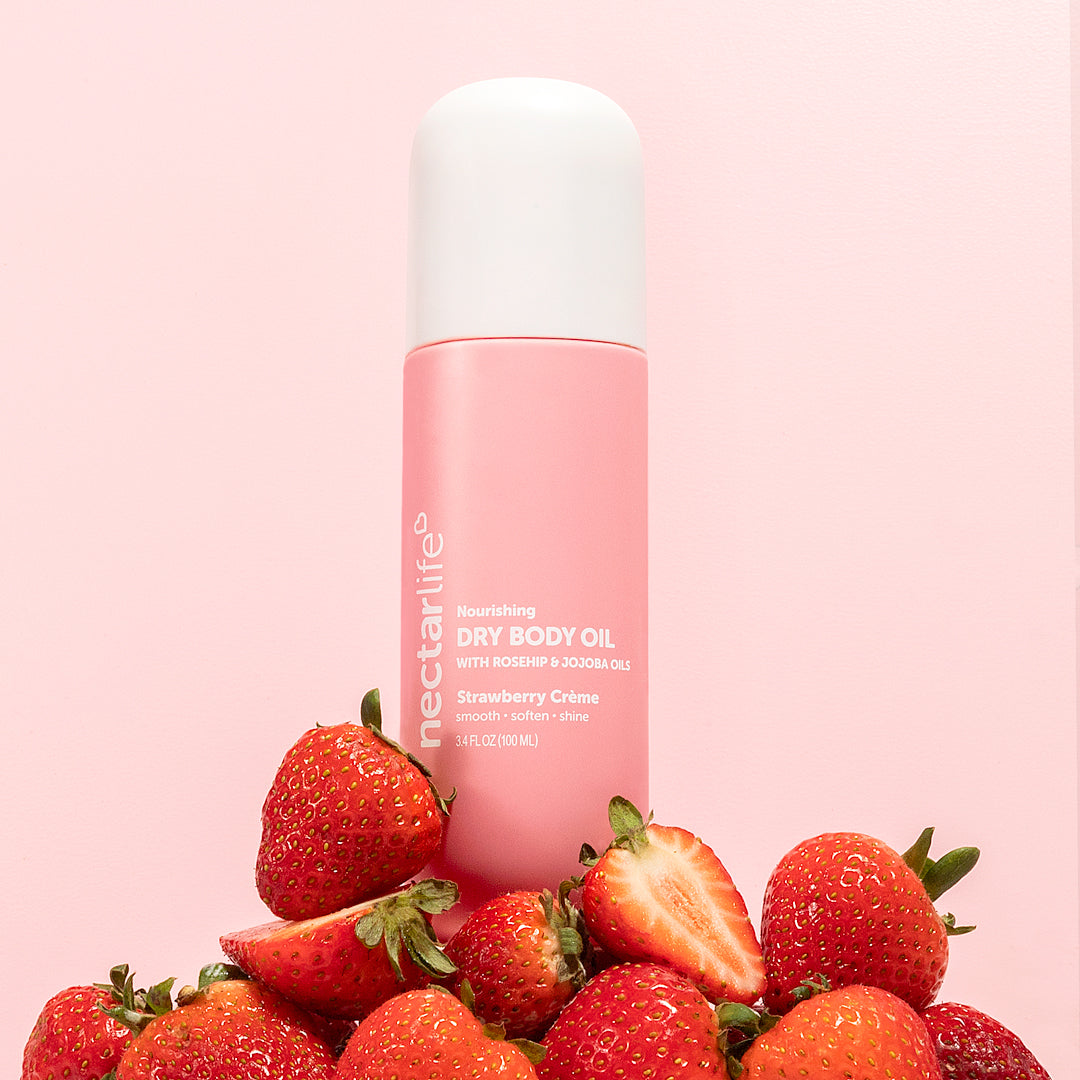 Strawberry Crème Nourishing Dry Body Oil by Nectar Life, pink spray bottle surrounded by fresh whole and sliced strawberries on a pink background, with rosehip oil and jojoba oil, 3.4 fl oz