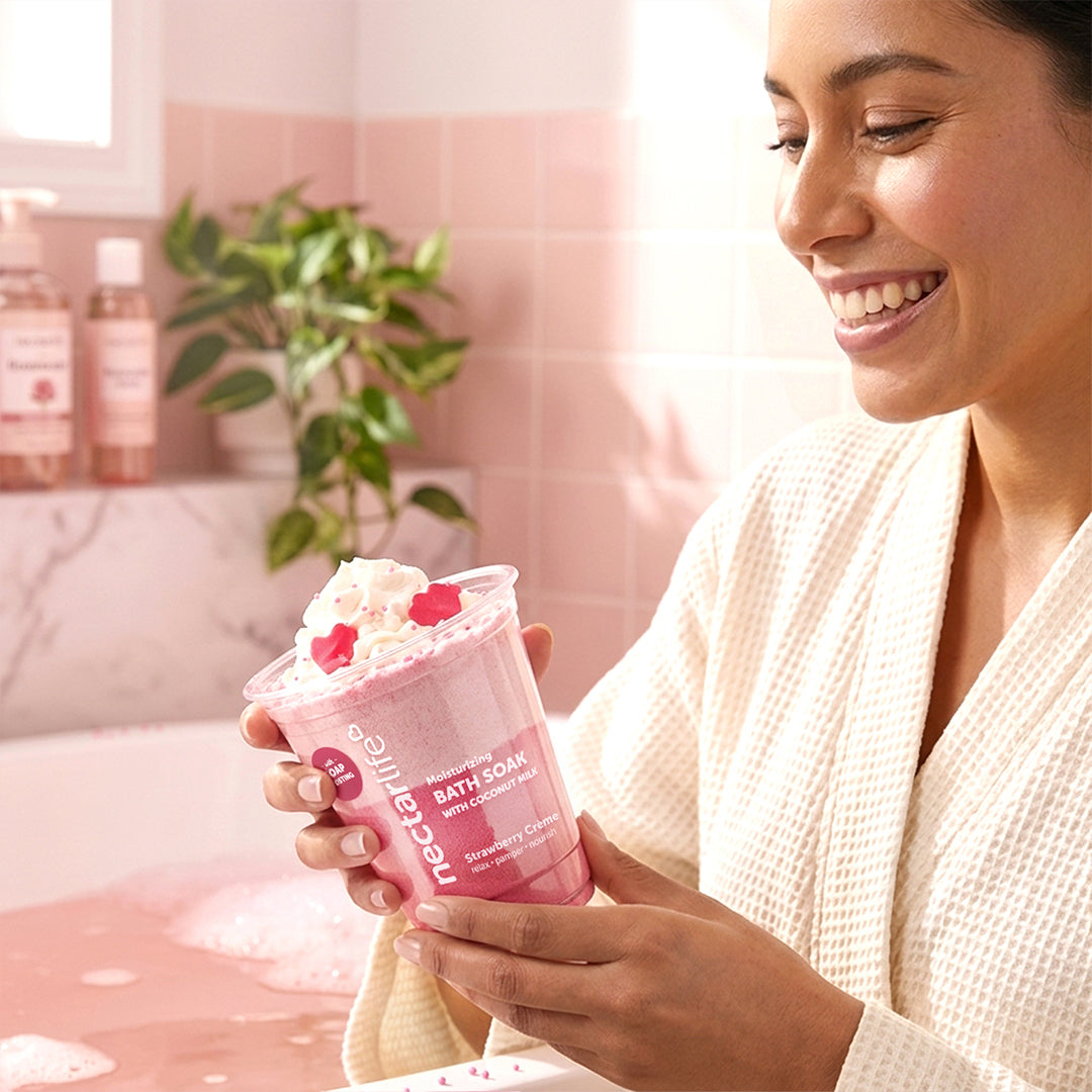 Woman smiling and holding Nectar Life Strawberry Crème Milkshake Bath Soak cup in a sunlit bathroom, fizzing bath soak with coconut milk, 9oz