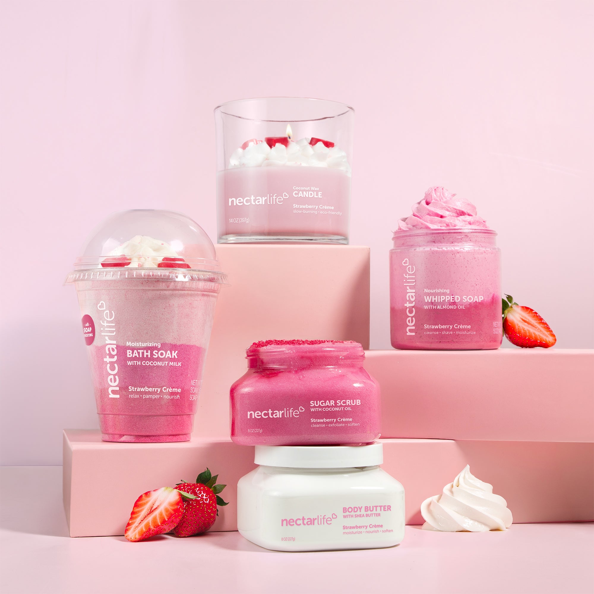 Nectar Life Strawberry Crème collection including Milkshake Bath Soak, Sugar Scrub, Whipped Soap, Body Butter, and Candle styled on pink pedestals with strawberries and whipped cream