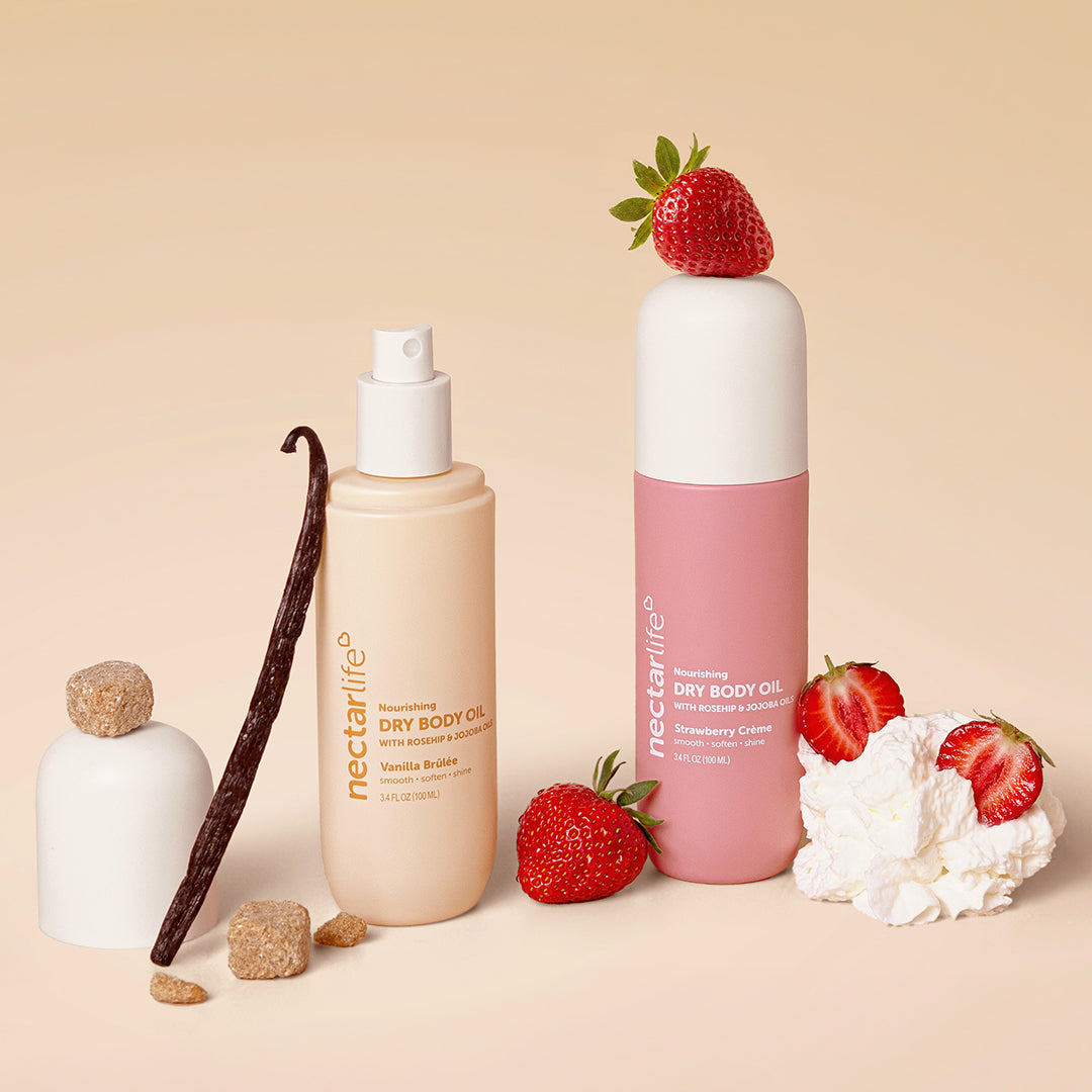 Nectar Life Nourishing Dry Body Oil in Vanilla Brûlée and Strawberry Crème, both 3.4 fl oz bottles styled with vanilla bean, brown sugar, fresh strawberries, and whipped cream on a warm neutral background