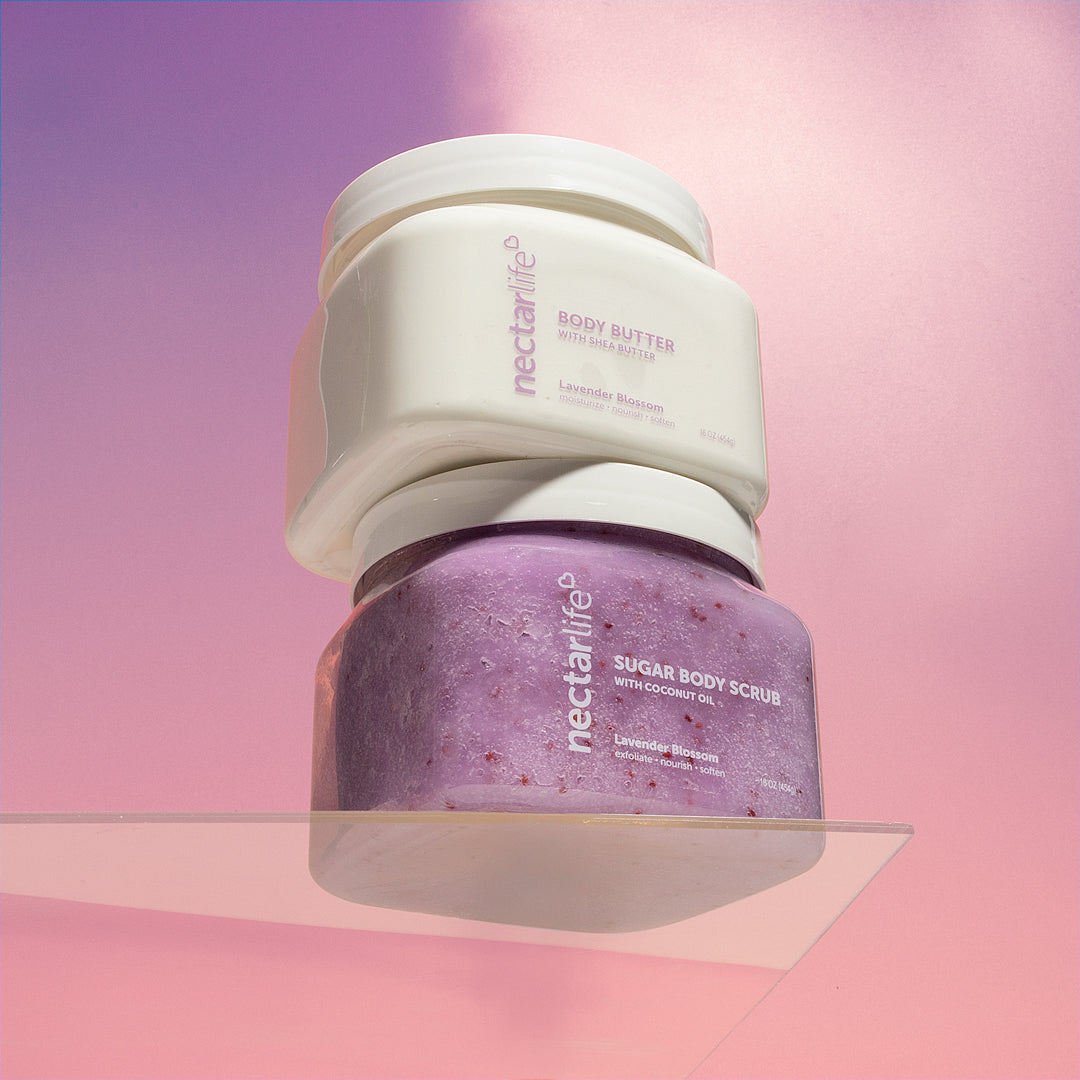 Product shot of Lavender Blossom Jumbo Scrub & Butter Duo by Nectar Life, 16oz Body Butter with Shea Butter stacked on 16oz Sugar Body Scrub with Coconut Oil on a clear acrylic shelf against a purple and pink background
