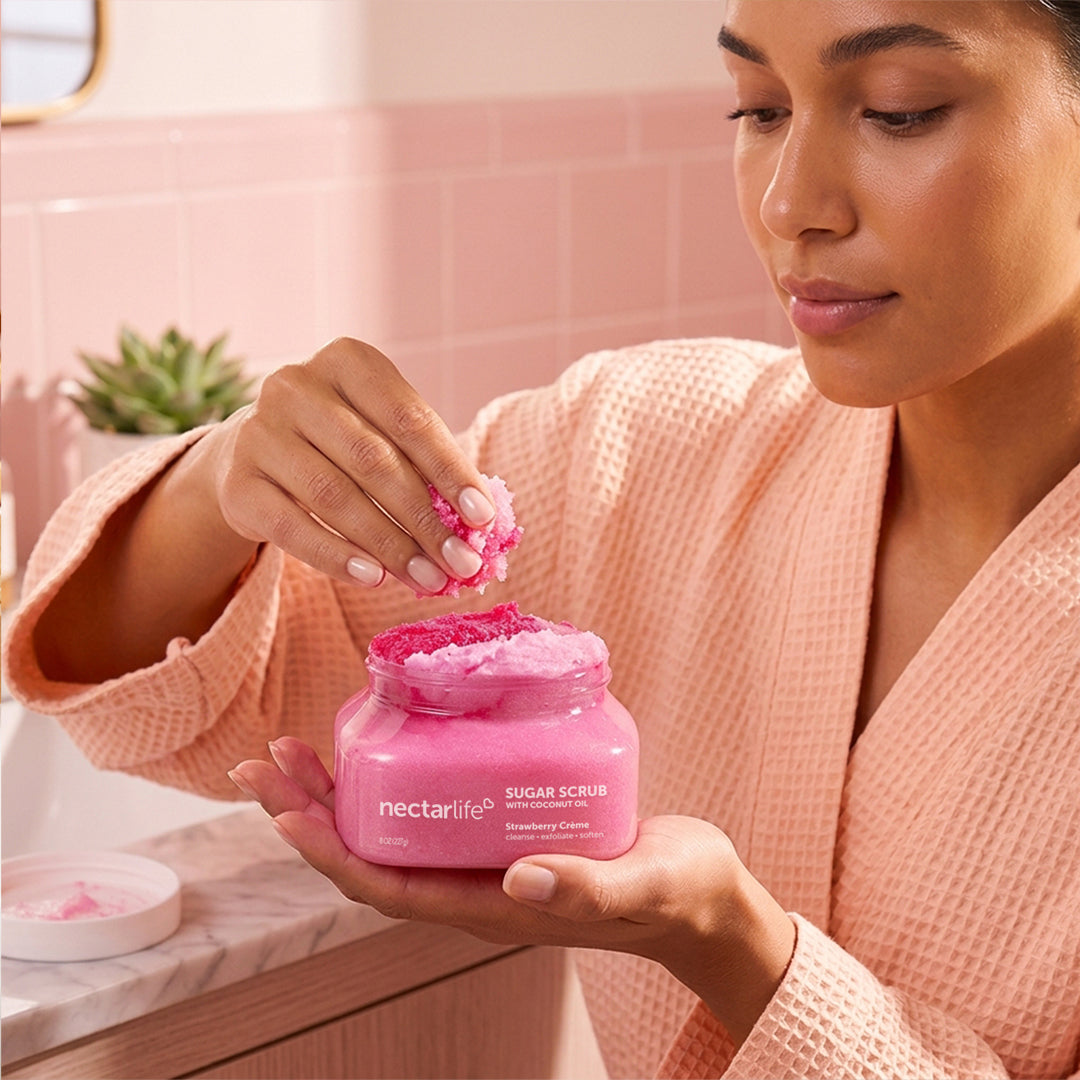 Woman scooping Nectar Life Strawberry Crème Sugar Body Scrub from jar in a pink bathroom, natural sugar exfoliants with shea butter and coconut oil