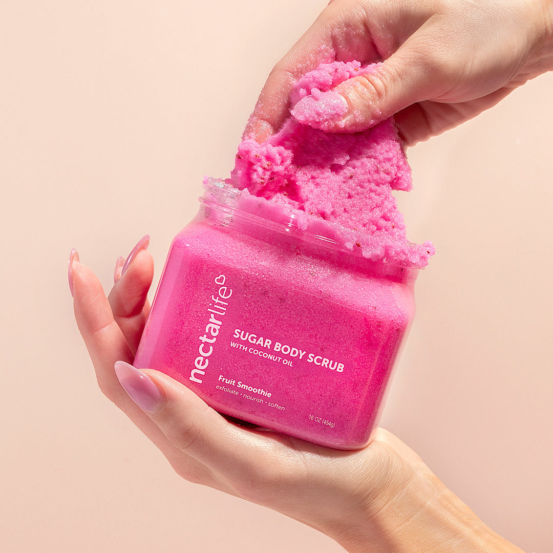 Hands holding an open jar of Nectar Life Fruit Smoothie Sugar Body Scrub with Coconut Oil 16oz with fingers scooping out vibrant pink scrub on a warm beige background