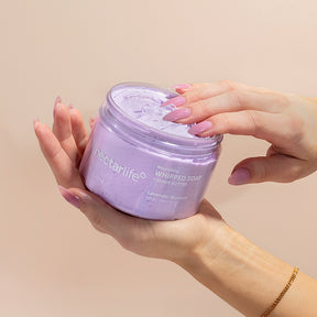 Hands cradling an open jar of Nectar Life Lavender Blossom Nourishing Whipped Soap and Shave Butter 10oz with pink manicured fingers dipping into the lavender whipped soap on a warm beige background