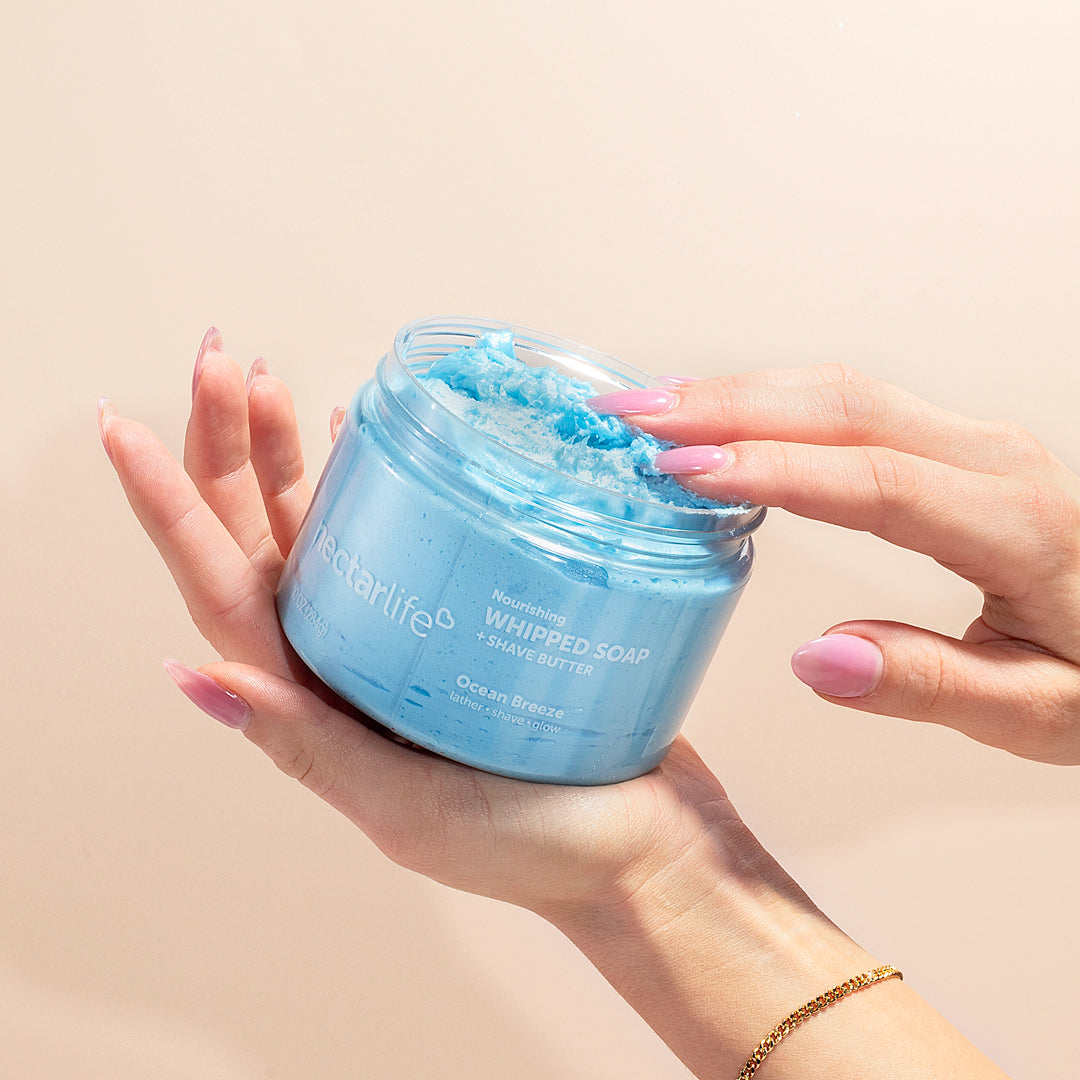 Hands cradling an open jar of Nectar Life Ocean Breeze Nourishing Whipped Soap and Shave Butter 10oz with pink manicured fingers dipping into the blue whipped soap on a warm beige background