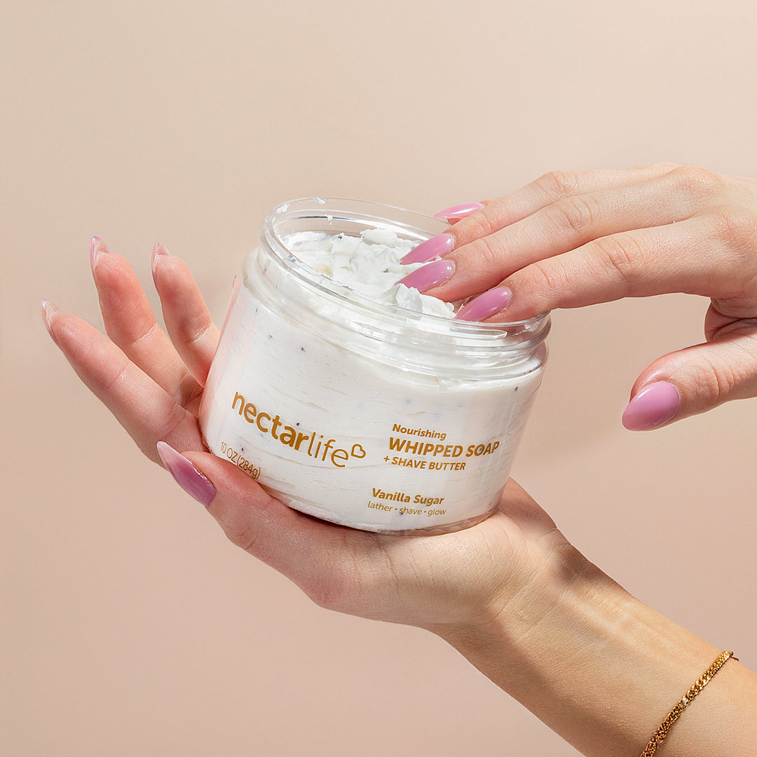 Hands cradling an open jar of Nectar Life Vanilla Sugar Nourishing Whipped Soap and Shave Butter 10oz with pink manicured fingers dipping into the white whipped soap on a warm beige background