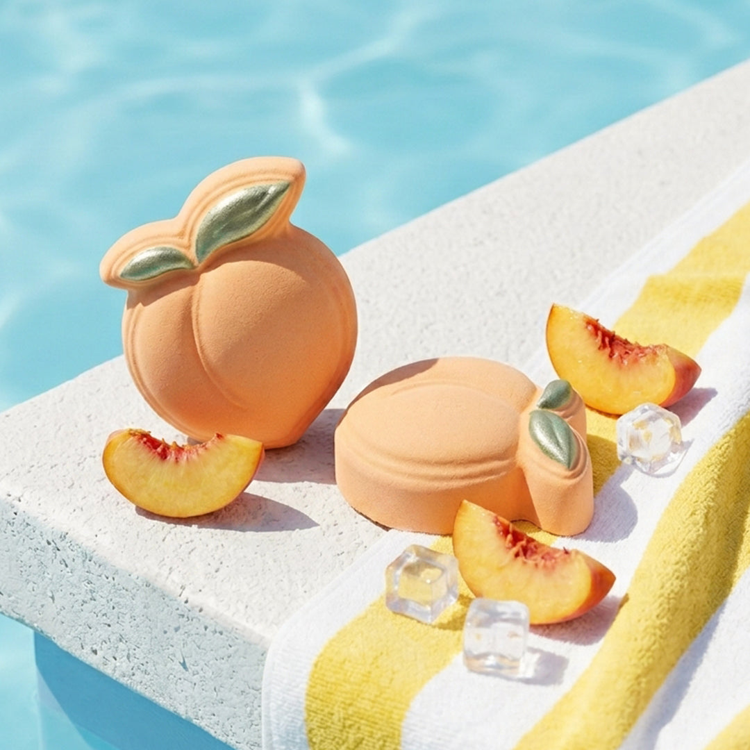 Nectar Life Juicy Peach Bath Bombs on a sunny pool ledge surrounded by fresh peaches and ice cubes, with a yellow striped towel and blue water in the background
