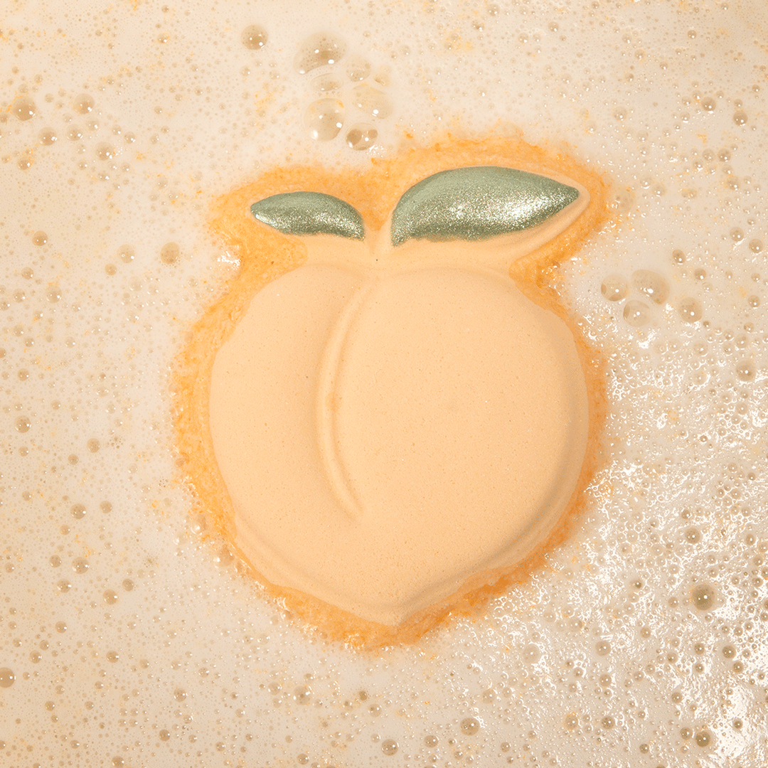 Nectar Life Juicy Peach Bath Bomb floating in warm fizzing bath water, peach-shaped bath bomb with glittery green leaf detail dissolving in a bubbly soak