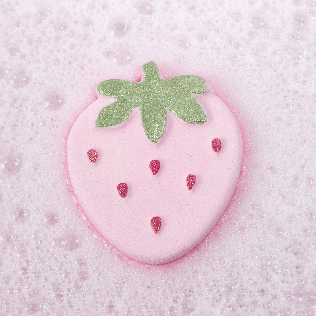 Nectar Life Strawberry Bliss Bath Bomb floating in pink fizzing bath water, showing the strawberry-shaped bath bomb with glittery green leaf top and red seed details dissolving in a bubbly soak