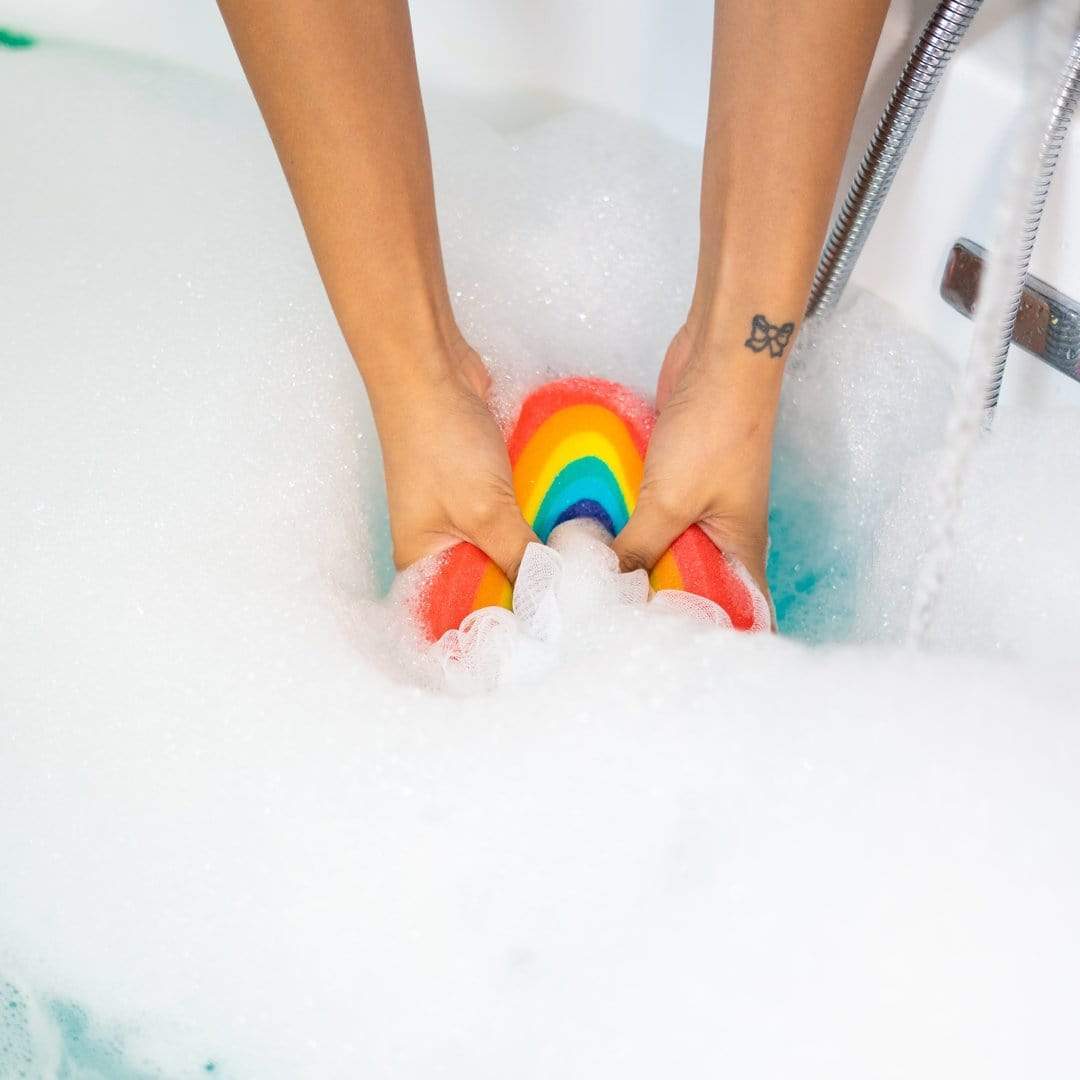 Hands squeezing the Nectar Life Large Rainbow Loofah Bath Sponge into a bubble-filled bathtub, showing the rainbow sponge body submerged in white foam with blue water visible at the edge