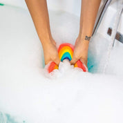 Hands squeezing the Nectar Life Large Rainbow Loofah Bath Sponge into a bubble-filled bathtub, showing the rainbow sponge body submerged in white foam with blue water visible at the edge