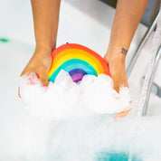 Hands holding Nectar Life Large Rainbow Loofah Bath Sponge over a sudsy bathtub, showing the colorful rainbow sponge lathering with white foam around the mesh loofah clouds